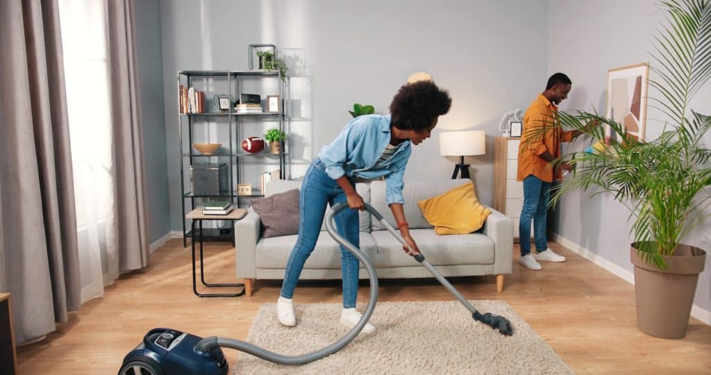 young nice happy married couple cleaning living room in apartment, wife vacuuming carpet floor while husband wiping dust on furniture behind, family concept, routine life