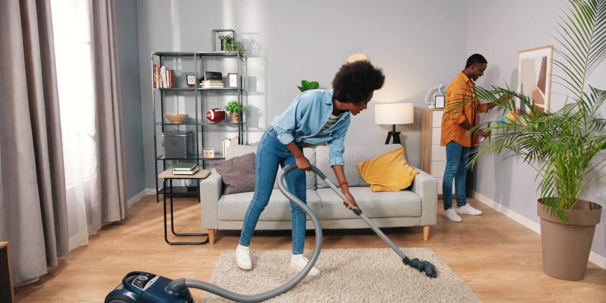 young nice happy married couple cleaning living room in apartment, wife vacuuming carpet floor while husband wiping dust on furniture behind, family concept, routine life